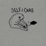 The self care shirt features hand written style "Self Care" with a stretching frog with a pickleball paddle and ball laying next to him. Grey shirt background.